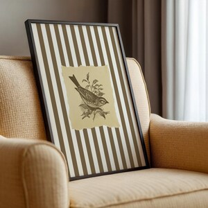 Op de afbeelding: Ingelijste kunst met een gedetailleerde vogelillustratie op een gescheurde papieren achtergrond. Het kunstwerk heeft een achtergrond van verticale bruine en witte strepen, in een zwart frame. Het kunstwerk leunt tegen een beige fauteuil.