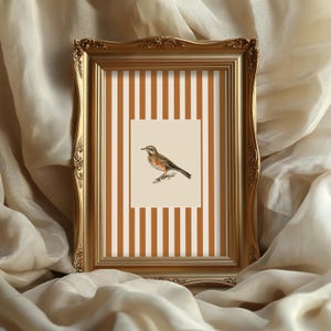 Könnte beinhalten: Ein gerahmter Druck mit einer detaillierten Illustration eines Vogels auf einem Ast. Das Kunstwerk befindet sich vor einem cremefarbenen Hintergrund mit braunen und weißen Streifen. Der Rahmen ist verziert und goldfarben, was eine Vintage-Ästhetik verleiht.