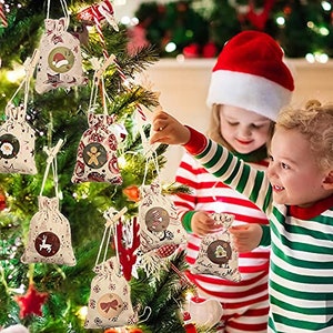 Könnte beinhalten: Ein Weihnachtsbaum, der mit Adventskalender-Säckchen aus Sackleinen mit verschiedenen Weihnachtsmotiven geschmückt ist. Die Säckchen hängen mit Schnur am Baum und zeigen verschiedene Motive, darunter ein Lebkuchenmann, ein Rentier und ein Schneemann. Zwei Kinder stehen in der Nähe des Baumes, eines hält ein Säckchen in der Hand, das andere schaut den Baum an.