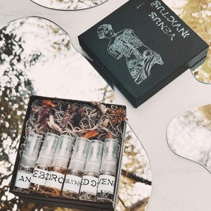 May include: A black box with the text "VENUS INVICTUS" and a skeletal illustration. Inside, a wooden box contains several small perfume bottles with labels. The bottles are filled with a light brown liquid and are surrounded by dried plants.