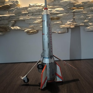 May include: A vintage silver toy rocket with red and blue accents, featuring the text "HOLDRAKETA" on its side. The rocket has a long, silver antenna and small wheels.