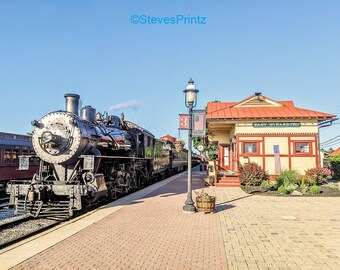 May include: A vintage steam train at the East Strasburg station. The black locomotive is positioned on tracks next to a red brick platform. The station building is yellow with a red roof. The sky is clear blue.