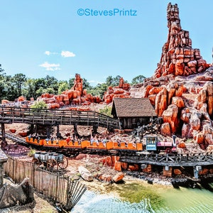 May include: An orange roller coaster train travels along a wooden track, passing a small wooden building and a rocky mountain formation. The scene is set against a bright blue sky, with the text "StevesPrintz" visible.