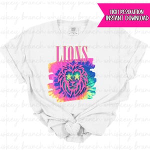 Może przedstawiać: Biała koszulka z wzorem tie-dye, różową grafiką lwa i napisem "LIONS" w kolorze różowym. Lew ma oczy w kształcie gwiazd. Koszulka jest marki Comfort Colors.