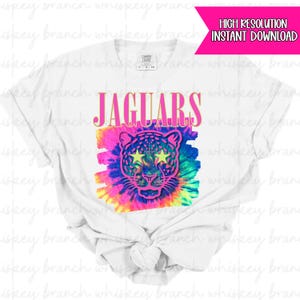 Może przedstawiać: Biała koszulka z wzorem tie-dye, twarzą jaguara i napisem "JAGUARS" w kolorze różowym i złotym. Jaguar ma oczy w kształcie gwiazd. Koszulka marki Comfort Colors.