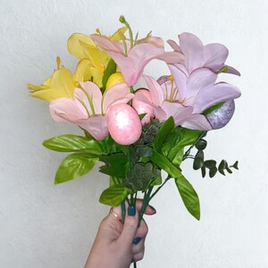 May include: A bouquet of artificial flowers with yellow, pink, and lilac lilies, green leaves, and Easter eggs. The arrangement is held in a hand with blue nail polish.
