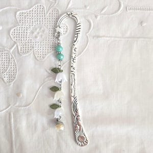 Peut inclure: Un marque-page en métal argenté avec un dessus incurvé et un motif floral décoratif. Le marque-page comporte un charm pendant avec des perles turquoise, des fleurs de muguet blanches et une petite perle transparente. Le marque-page est posé sur un fond blanc texturé.