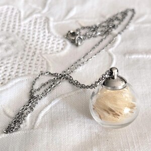 Peut inclure: Un collier argenté avec un pendentif en forme de globe de verre transparent rempli d'une matière plumeuse de couleur claire. Le pendentif est surmonté d'un capuchon en métal argenté et est suspendu à une chaîne délicate.