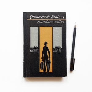 Könnte beinhalten: Ein Vintage-Buch mit schwarzem Einband, auf dem der Titel "Gunteris de Broinas Buridano asilas" in Weiß steht. Das Cover-Design zeigt eine Silhouette einer Person mit Gepäck vor einem goldenen und grauen Hintergrund. Ein schwarzer Stift liegt neben dem Buch.
