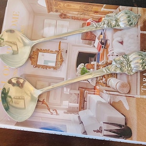 May include: Two silver serving spoons with ornate floral designs on the handles. One spoon has a slotted bowl, and the other has a round bowl. The spoons are resting on a book with the title "TARA SHAW COLLECTION".