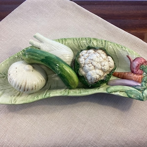 May include: A ceramic vegetable-shaped serving dish. The dish is leaf-shaped and holds a garlic bulb, a cucumber, a head of cauliflower, carrots, and other vegetables. The dish is painted in naturalistic colors.