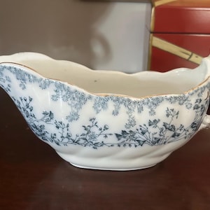 May include: A white ceramic gravy boat featuring a blue floral pattern around the rim and body. The vessel has a scalloped edge, a curved handle, and a pouring spout. The rim and handle display visible signs of wear with faded gold-tone trim.