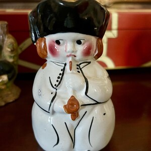 Puede incluir: Figura de cerámica de una persona con abrigo blanco y sombrero negro. La figura tiene mejillas rosadas, cabello castaño y una expresión severa. El abrigo tiene ribetes negros y la figura sostiene un objeto marrón.