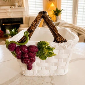 Peut inclure: Un panier en céramique blanche avec une texture tressée et une anse marron en forme de branche d'arbre. L'avant est orné d'une grappe de raisins violets brillants et d'une feuille verte attachée à une vigne, créant un relief décoratif.