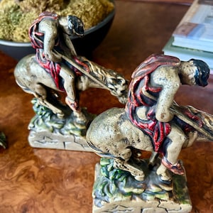 May include: Two antique figurines depicting riders on horseback. The figures are painted in gold, red, and black, with detailed features. The bases are green and brown, suggesting a grassy terrain. The figurines are displayed on a wooden surface.