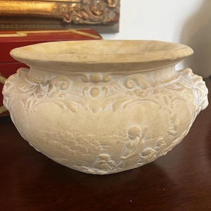 May include: A beige, antique-style planter with ornate relief carvings. The planter features a wide rim and two small handles. The relief depicts figures in a landscape. The overall design is reminiscent of classical art.