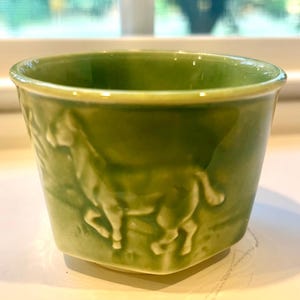 Peut inclure: Une petite tasse en céramique vert clair présentant un motif en relief d'un cheval au galop sur son extérieur. La tasse possède une finition lisse et brillante avec des variations subtiles dans la glaçure verte, créant un aspect monochrome.