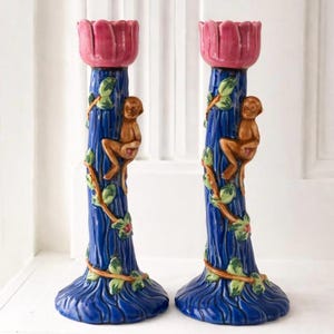 May include: Pair of ceramic candle holders. Each features a blue tree trunk design with a brown monkey climbing the side. The top is a pink flower shape. Green leaves and vines wrap around the trunk.