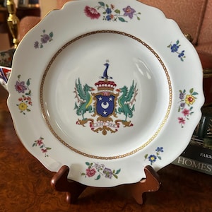 Puede incluir: Un plato de porcelana blanca con borde festoneado y ribete dorado. El centro presenta un escudo heráldico colorido con dos dragones verdes, un escudo azul con una luna creciente y estrellas blancas, y el texto LAUS DEO. Flores decoran el borde.