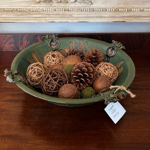 May include: A round, dark green ceramic bowl with decorative leaf-shaped handles, filled with a collection of natural items including brown pinecones, woven rattan spheres, smooth wooden balls, and small green moss-like textured spheres.