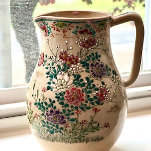 Puede incluir: Jarra de cerámica color crema con asa beige y diseño floral. La jarra presenta flores rojas, moradas y blancas con hojas y tallos verdes. El borde tiene un borde verde y rojo.