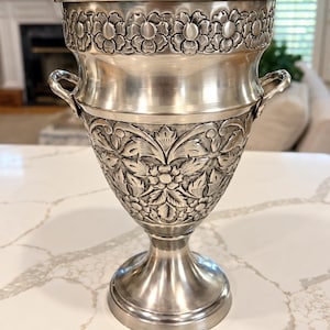 May include: A silver-toned metal wine cooler or vase featuring a flared rim with a repeating embossed floral band. The body displays intricate relief carvings of leaves and flowers, complemented by two small side handles and a pedestal base.
