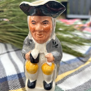 May include: A ceramic figurine of a man in a tricorn hat and colonial attire. The figure has a tan face, gray coat, and black shoes. It holds two yellow and black objects, possibly lanterns or jugs. The figurine is on a plaid surface.