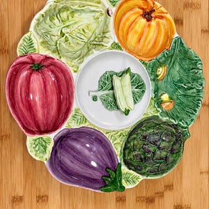 May include: A round ceramic serving platter featuring a central white bowl with a small green vegetable garnish. The platter is divided into sections shaped like a red tomato, a purple eggplant, a green artichoke, a pumpkin, and cabbage leaves.