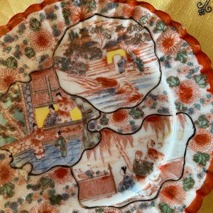 May include: A white porcelain plate with a scalloped edge and a red rim. The plate is decorated with a floral pattern in shades of red, green, and blue. The center of the plate features three scenes of Japanese figures in a landscape setting.