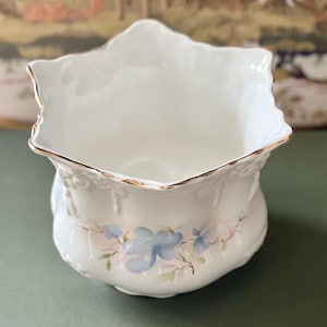 Könnte beinhalten: Ein weißer Porzellanblumentopf mit gewelltem Rand und goldenem Besatz. Der Topf hat ein Blumendesign mit blauen und rosa Blüten. Die Außenseite hat ein strukturiertes, geprägtes Muster.