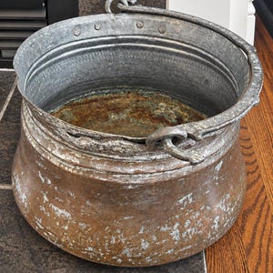 May include: A large, round, weathered copper pot with a wide opening and a thick, textured rim. The exterior shows signs of oxidation and wear, while the interior base features a prominent layer of rust. It includes a sturdy metal handle.
