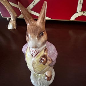 May include: A ceramic figurine of a rabbit wearing a purple jacket and yellow vest. The rabbit has brown ears and is standing on a small patch of green grass. The figurine is a decorative item.