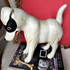 May include: A white dog figurine with a black mask around one eye and a bone-shaped object. The dog is standing on a stack of books with the titles "Enduring Southern Homes" and "Eric Ross". The background includes a red pillow.
