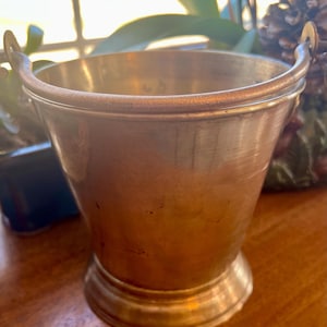 Puede incluir: Un pequeño cubo de latón antiguo con dos asas pequeñas. El cubo tiene forma acampanada y base redondeada. El latón tiene una pátina cálida y envejecida. El cubo es probablemente decorativo.