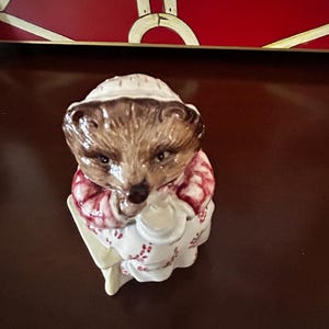 以下が含まれることがあります： 白い帽子と赤と白の模様のドレスを着たハリネズミの陶器製フィギュア。ハリネズミは白いものを持っています。フィギュアは茶色とタン色の毛皮で、茶色の表面に座っています。