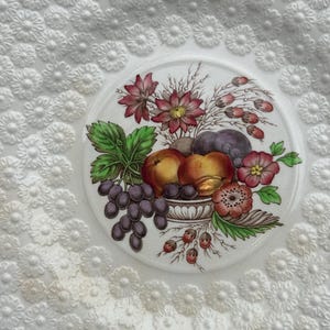 May include: White ceramic plate with embossed floral pattern. The center features a detailed illustration of fruit and flowers in a decorative bowl. The plate's edge has a scalloped design.