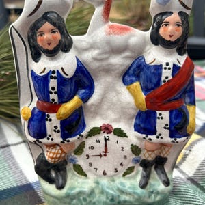 May include: A vintage ceramic clock featuring two figures in blue coats and hats, with yellow gloves and red sashes. A white deer with red spots is in the center. The clock face shows the time as 12:00.
