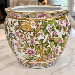 Könnte beinhalten: Ein weißer Keramik-Fischschalen-Übertopf mit einem farbenfrohen, handbemalten botanischen Muster aus grünen Blättern, rosa und blauen Blüten sowie goldenen Akzenten. Das runde Gefäß hat einen breiten, ausgestellten Rand und eine glatte Oberfläche.