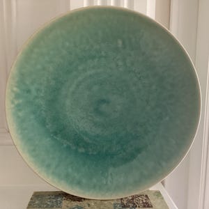 Puede incluir: Un plato de cerámica grande y redondo con un esmalte color turquesa. El plato tiene un borde ligeramente elevado y una superficie texturizada. El color es una mezcla de turquesa y blanco, creando un efecto veteado. El plato es adecuado para servir alimentos.