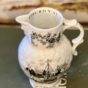 May include: A white ceramic jug with a spout and handle. The jug features black floral and landscape designs, with the text "1751 ROYAL" around the rim. The base has additional text. The jug has a bulbous body.