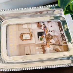 Puede incluir: Una bandeja rectangular plateada con borde de cuentas y asas curvas. La bandeja contiene una imagen enmarcada con el texto "COLOR OF THE BOTTS" y "TARA SHAW". La imagen representa un interior de habitación.