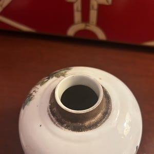 Pu&ograve; includere: Un vaso rotondo in ceramica bianca con un'apertura scura e un bordo marrone. Il vaso presenta dettagli dipinti in verde e nero sbiaditi. Lo sfondo include una scatola decorativa rossa.