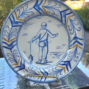 Puede incluir: Un plato de cerámica redondo con un borde decorativo pintado a mano en azul y dorado, que presenta una ilustración central de una figura de pie sosteniendo una pala. El diseño está realizado en tonos azules y dorados sobre un fondo crema.