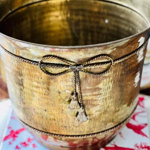 May include: A cylindrical brass planter featuring a hammered texture on the exterior surface. The vessel is decorated with a raised rope-like trim encircling the middle, forming a bow detail with two small hanging tassels attached to the centre.