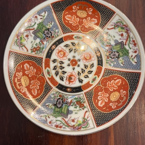 May include: A decorative ceramic plate with a detailed floral and geometric design. The plate features a central floral motif surrounded by alternating red and black panels with intricate patterns. The rim is white with gold accents.