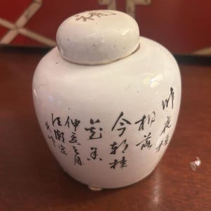Pu&ograve; includere: Un piccolo vaso di ceramica bianca con corpo arrotondato e coperchio. Il vaso &egrave; decorato con calligrafia nera. Il coperchio presenta un design semplice. Il vaso &egrave; su una superficie di legno.