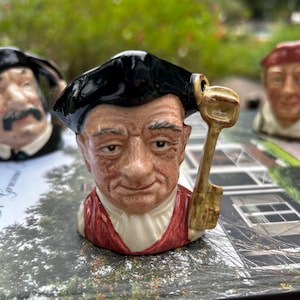 May include: A ceramic character jug depicting a person wearing a black tricorn hat and a red coat with a white cravat. The figure holds a large, tan-coloured key in the right hand. The facial features include detailed wrinkles and a neutral expression.