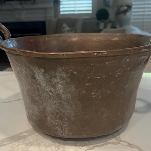 May include: A large, round, weathered copper basin featuring a wide, open top and two sturdy, curved metal handles attached to opposite sides. The exterior surface displays a dull, brownish-metallic patina with visible signs of oxidation and wear.