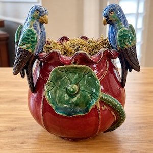 May include: A round, glossy red ceramic planter with a ruffled rim, featuring two blue and green parrot figurines perched on opposite sides and a large, sculpted green leaf relief on the front. The interior is filled with dried green moss.
