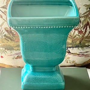 Puede incluir: Una jardinera de cerámica turquesa con una parte superior rectangular y una base de pedestal. La jardinera tiene un acabado craquelado y una fila de pequeños puntos blancos alrededor del borde. La base es cuadrada.